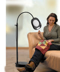 Go-To LED Floor Lamp And Table Lamp With 5-Inch Magnifier next to woman knitting on sofa, sleek black design, flexible arm