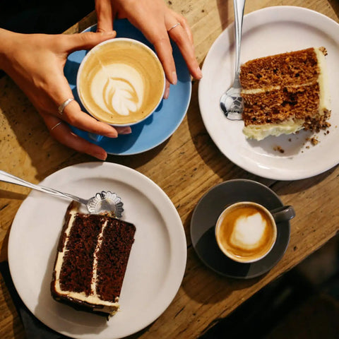 Top view of two plates with cake slices and cappuccinos on a wooden table, one served with a floral spoon.