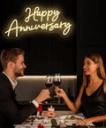 Couple toasting under 'Happy Anniversary' neon sign during a romantic dinner.