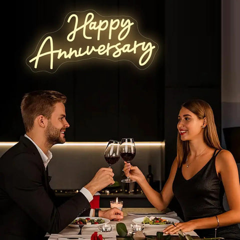 Couple toasting under 'Happy Anniversary' neon sign during a romantic dinner.