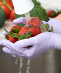 Person washing strawberries wearing Lavender Nitrile Exam Gloves 3.1 Mil 9.5 Inches Medium All Day Comfort Box Of 250 Powder Free Non Sterile Disposable Gloves With Low Dermatitis Potential And Chemical Resistance For Fentanyl And Gastric Acid Per ASTM D6978.