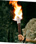 Person holding a flaming wood torch, focus on glowing flame, for Outdoor Wood Torch Lantern For Garden And Patio Decor, Flickering Flame Light For Walkways And Yards, Rattan Style Backyard Tiki Torch, Vintage Brown Wood Lantern For Parties, Campouts, And Cozy Lawn Vibe.