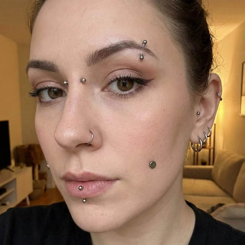Person with multiple facial and ear piercings featuring nose, lip, and eyebrow jewelry in a living room setting.