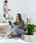 Woman sitting on floor holding photo frame with potted plant in storage box next to her; man organizes items in background