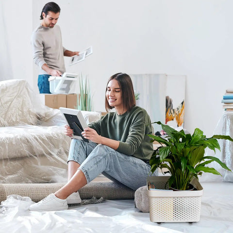 Woman sitting on floor holding photo frame with potted plant in storage box next to her; man organizes items in background