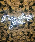 White neon sign reading 'It was always you' against a floral wall backdrop with white roses.