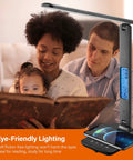 LED Desk Lamp With Wireless Charger And USB Port beside a family reading, showing clock, alarm, date, and temperature display
