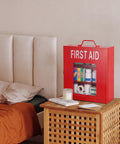 Red first aid box with visible supplies, placed on a wooden table beside a bed.