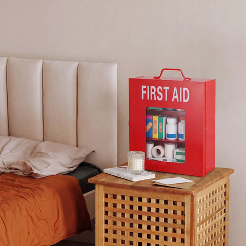 Red first aid box with visible supplies, placed on a wooden table beside a bed.