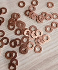 Solid Annealed Pure Copper Flat Washers Kit Vibrant Orange M5 To M20 With 1Mm And 2Mm Thickness Ideal For Hardcore Auto Repair Where You Need Reliable Sealing Performance On Heavy Load Application Without Struggling Too Much At All. copper washers spread on a wood surface