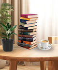 Stack of books, cup of coffee, and plant on a wooden table in a cozy room with curtains in the background.