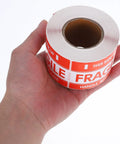 Person holding roll of Fragile Red Shipping Labels With High Visibility And Strong Adhesive, Paper Warning Stickers On Rolls For Small Business Packaging