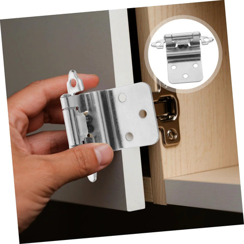 Front view of a silver self-closing inset cabinet hinge held by hand beside a light wood cabinet door