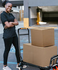 Folding hand truck with black steel frame and orange handle, beside two cardboard boxes in a parking area