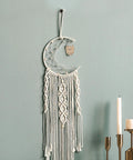 Crescent moon macrame wall hanging with owl design and tassels next to candles on a light green wall.
