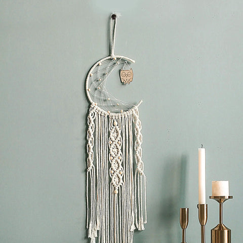 Crescent moon macrame wall hanging with owl design and tassels next to candles on a light green wall.