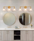 Three Light Gold Vanity Light Fixtures With White Frosted Glass Shades Over Mirror In Brushed Antique Gold Finish above bathroom sinks