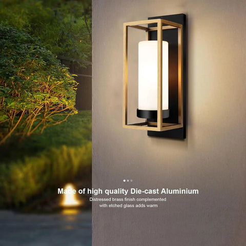 Close-up of a modern black and gold outdoor wall sconce with white diffuser installed on a wall, showcasing brass accents at night.