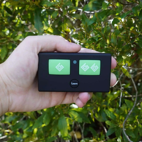 Person holding AXR1 Water-Resistant 3-Button Remote For Automatic Gate Openers against greenery background.