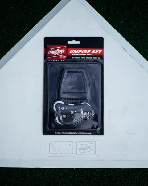 Umpire Gear Kit With 4-Dial Indicator, Plate Brush, And Pro-Style Cloth Bag packaged on a baseball home plate background.