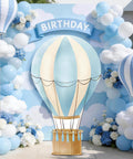 Blue Hot Air Balloon Cardboard Cutout For Baby Shower And Birthday Party Photo Booth Backdrop Life Size Standup Prop With Easy Setup Includes Sticks Tape And Backboard For Party Decor set among balloons and flowers.