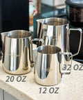 Display of 32 Oz Milk Frother Pitcher with Clear Scale, alongside 20 Oz and 12 Oz stainless steel jugs on a kitchen counter.