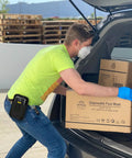 Man loading car with boxes of disposable face masks, wearing a mask and gloves, scanner holster attached to belt.