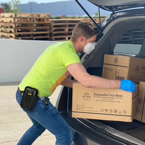 Man loading car with boxes of disposable face masks, wearing a mask and gloves, scanner holster attached to belt.