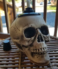 Close-up of a skull-shaped tiki torch on a wicker table, part of a Two Pack Skull Tiki Torch Outdoor Patio Decor For Halloween And Gothic Vibes, Epic Fire Display Skull Table Torches To Elevate Your Garden And Patio Lighting, Durable Outdoor Flame Lighting, Lit.