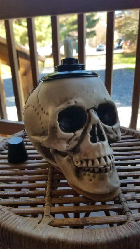 Close-up of a skull-shaped tiki torch on a wicker table, part of a Two Pack Skull Tiki Torch Outdoor Patio Decor For Halloween And Gothic Vibes, Epic Fire Display Skull Table Torches To Elevate Your Garden And Patio Lighting, Durable Outdoor Flame Lighting, Lit.