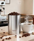Top Shelf Automatic Vacuum Coffee Canister With Stainless Steel Build Digital Display One Touch Freshness Seal Keeps Your Beans Fire And Flavor Loaded Extra Long Standby Battery Life filled with roasted coffee beans next to a steaming cup