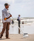 Heavy Duty Stainless Steel Sand Scoop For Metal Detecting With Long Handle Kit Includes Pinpointer Shovel And Gloves Beach Treasure Hunting Gear For Relics And Coins being used on a beach by a metal detector enthusiast.