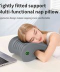 Woman resting on ergonomic gray nap pillow by desk, providing neck and head support.