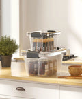 Stackable Plastic Storage Bins Set Clear 6 Quart Nestable Boxes With White Lids And Secure Latching Buckles 12 Pack Perfect For Pantry Toys Office And Garage Organization shown stacked on a kitchen counter containing bottles.