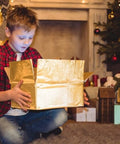 Child opening a golden box in a cozy, festive living room with Christmas decorations and a fireplace.