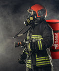 Firefighter wearing safety gear and holding equipment with fire suppression tools, standing against smoky background.