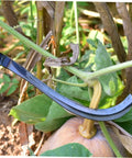 Close-up of a 2-Pack Mowing Sickle Weeding Hook with No Slip Grip cutting a plant stem in a garden setting, rustproof design visible.