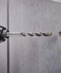 Installed SDS‑Plus masonry drill bit (12 mm) in a hammer drill drilling into concrete wall.