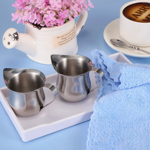 Two 3oz stainless steel creamer pitchers with handles on a tray, surrounded by a towel and decorative items. Product focus: 4 Pack 3oz Stainless Steel Creamer Pitchers With Handle For Coffee Milk Sauces.