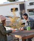 Two people sitting at a table outside an RV, playing cards with a green mug, autumn trees in the background.