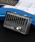 10 Band EQ Pedal for Electric Guitar and Bass With 31.25 Hz to 16 KHz Range, Each Band Delivers Up to 12 dB Gain or Cut, Durable Aluminum Alloy Build, Easy Stage and Studio Tone Shaping for Electric Players on black surface next to blue electric guitar.