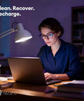 Person working at a laptop in a dimly lit room with text 'Clean. Recover. Recharge.' on the wall.