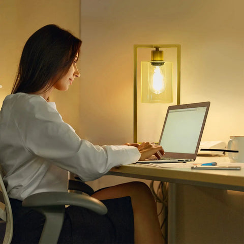 Industrial Table Lamp Set Of 2 With Gold Finish, clear glass shade emitting warm light on desk, woman working on laptop nearby