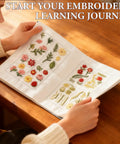 Person browsing Embroidery Stitch Book Kit With 106 Patterns, linen pages open displaying floral designs on wooden table