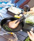 Cooking steak in Travel Ready 13 Piece Nonstick Cookware Set with lightweight cast iron pan and nylon tongs on campsite table