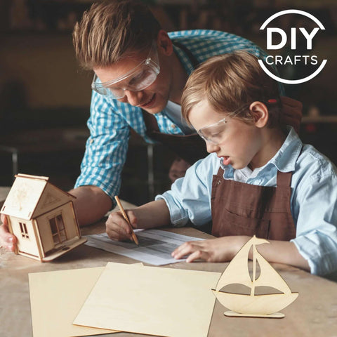 Front view: DIY Crafts basswood craft sheets on table with wooden house and sailboat; adult and child wearing safety goggles