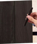 Hand holding a black stick over wooden surface, part of a 2 Pack Wood Filler Sticks for Furniture Repair Scratch Remover Pens Quick Fix Paint Markers for Desks Tables Floors Cabinets Bookcases Easy DIY Wood Repair Kit.