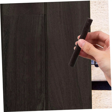 Hand holding a black stick over wooden surface, part of a 2 Pack Wood Filler Sticks for Furniture Repair Scratch Remover Pens Quick Fix Paint Markers for Desks Tables Floors Cabinets Bookcases Easy DIY Wood Repair Kit.