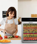 Top Shelf Heavy Duty Food Dehydrator With 12 Sleek Stainless Steel Trays on kitchen counter with sliced vegetables and fruits.