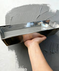 Hand holding shiny metal mudding tray filled with wall joint compound, used for wall and ceiling repairs in Kick Ass 11 Piece Drywall Tool Set With Telescopic Extension Pole And Paint Roller For Wall And Ceiling Mudding Taping Smooth Repairs And Home Renovation Work.